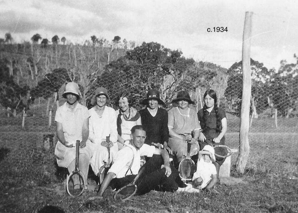 A tennis group of women and one man in front of a wire-netting court, c. 1934